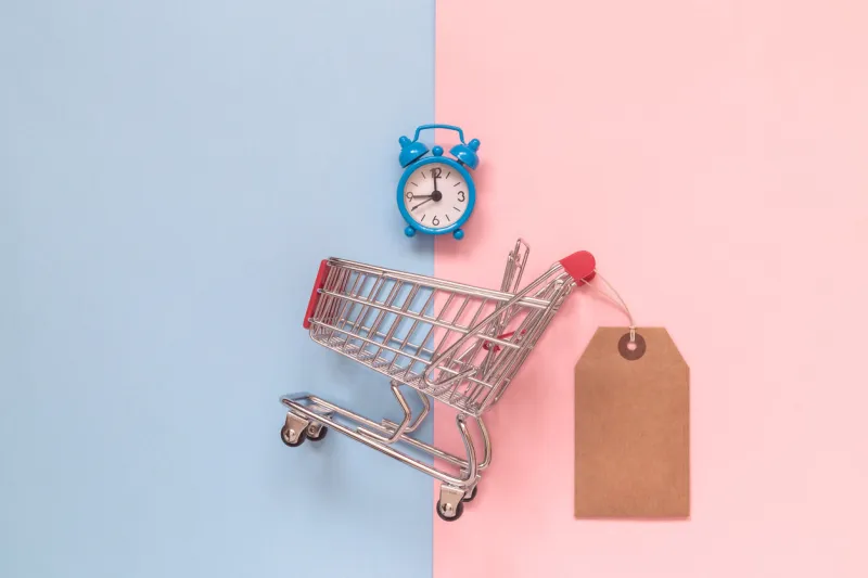shopping trolley toy with special offer tag and blue alarm clock on pastel background minimalistic flat lay concept