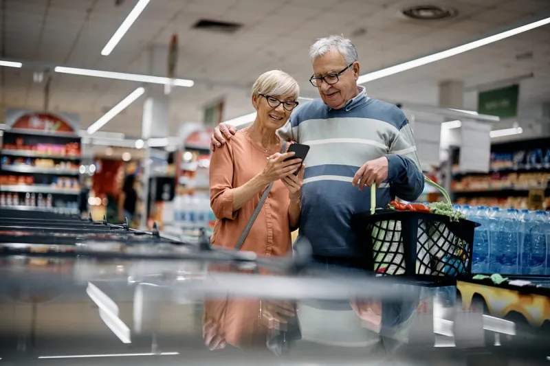 happy senior woman and her husband using app on mobile phone while shopping in supermarket