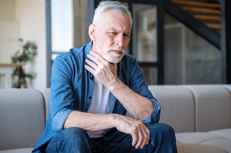 middle aged man suffering sore throat, touching neck and feeling sick while sitting alone on couch in living room unhappy frowning male has viral, infection or flu symptoms