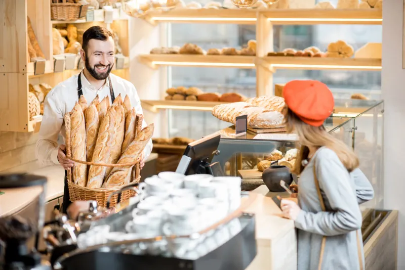 handsome bread seller with basket full of baguettes and female customer in the beautiful store with bakery products