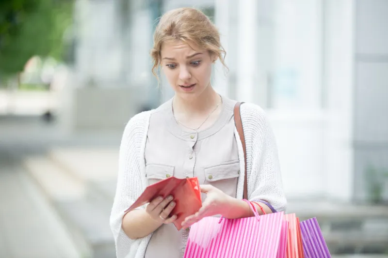 portrait of unhappy stressed beautiful person looking in open wallet with shocked expression while holding color shopping bags at mall entrance young model spent too much money during shopping time
