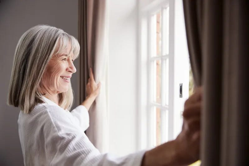 senior woman opening bedroom curtains and looking out of window