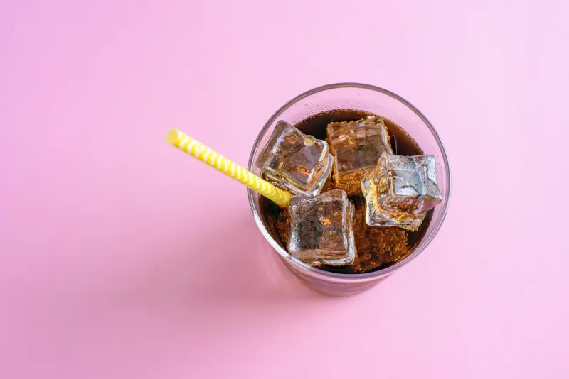 top view of a glass of cola with ice cubes and paper drinking straw on pink background