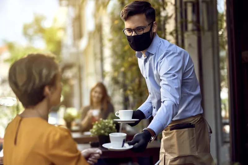 waiter wearing protective face mask while serving coffee to his guest at outdoor cafe