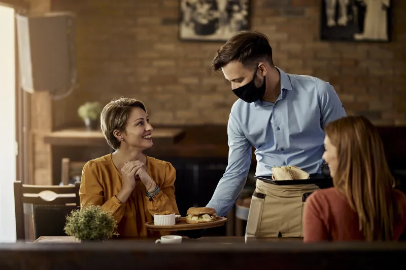 young waiter wearing protective face mask while serving food to his guests in a restaurant