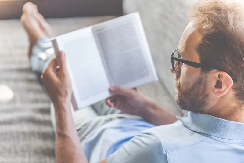 back view of handsome young businessman in casual clothes and eyeglasses reading a book while lying on couch at home