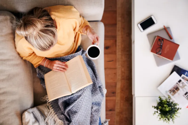 overhead shot looking down on woman at home lying on reading book and drinking coffee