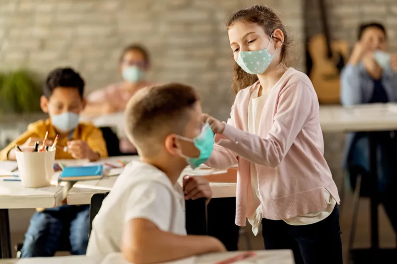 happy schoolgirl helping her classmate to properly put on a face mask while being at school during coronavirus epidemic