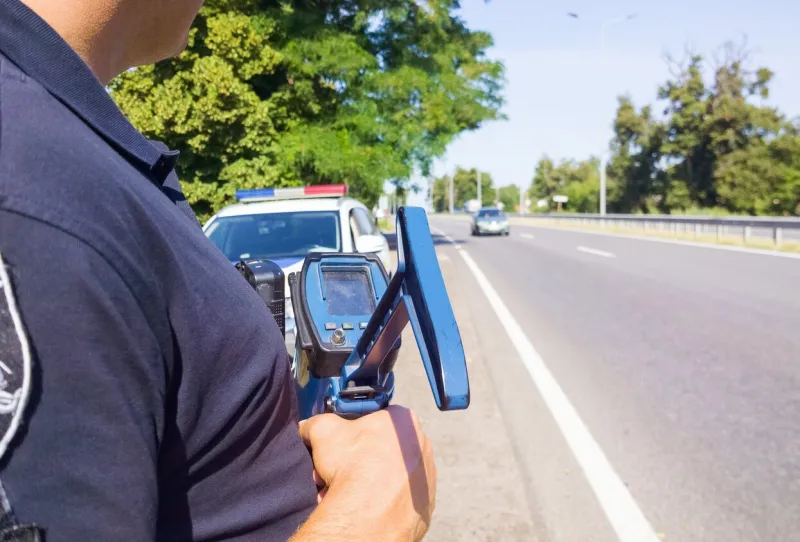 police officer holding laser speed gun near police car on highway background selective focus, part of body