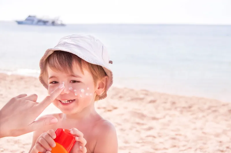 sun protection hand of caucasian mother applying suncream (suntan lotion) from a plastic container to her happy cute son before tanning during summer holiday on beach summer vacations concept copyspace, close up