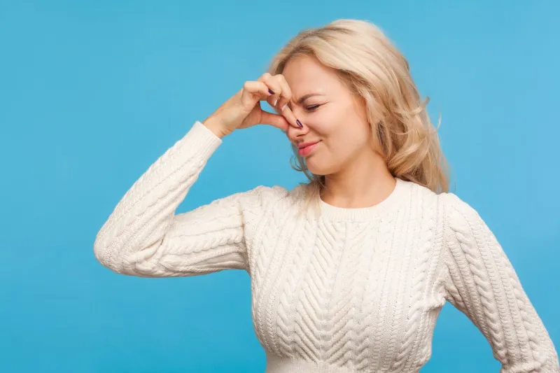 blond woman holding breath with fingers on nose, feeling unpleasant smell, disgusted odor, harmful emission indoor studio shot isolated on blue background