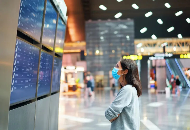 woman in virus protection face mask looking at information board checking her flight in international airport departure board, flight status