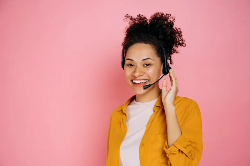 happy african american curly haired young woman with headset, help desk worker, call center operator, consultant, talking with client, stand on isolated pink background, looks at camera,smile friendly