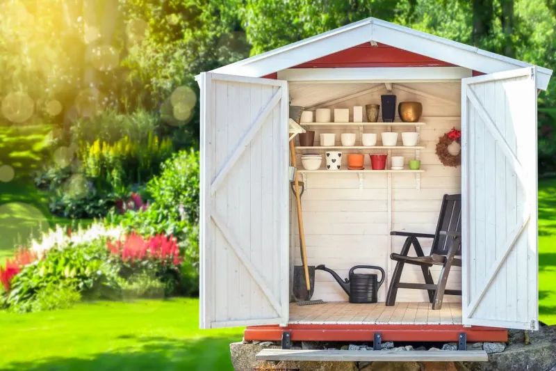 garden shed filled with gardening tools shovels, rake, pots, water pitcher in storage hut green sunny garden in the background