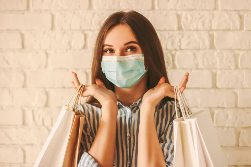 beautiful girl shopper in medical face mask holding shopping bags in hands young happy woman customer in protective mask on face with paper bags girl shopaholic coronavirus covid-19, sale, discount