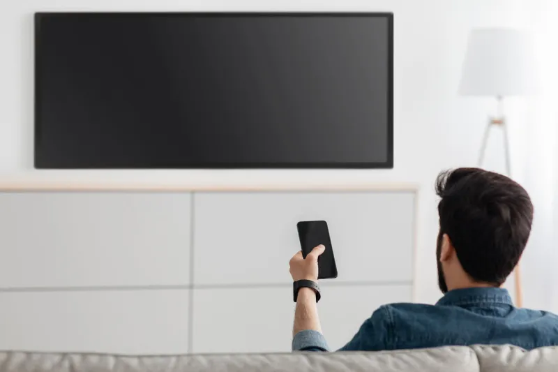 young man watching television with blank screen and using smart tv remote control application on smartphone, back view select movie stream, change channel or settings in the menu