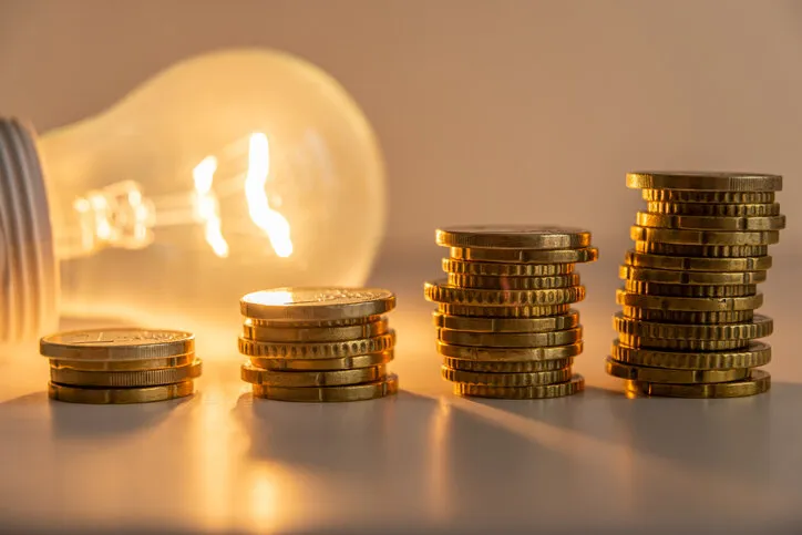 lit light bulb with coins beside it increase in energy tariffs efficiency and energy saving