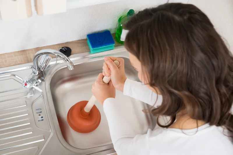 happy woman using plunger in kitchen sink at home