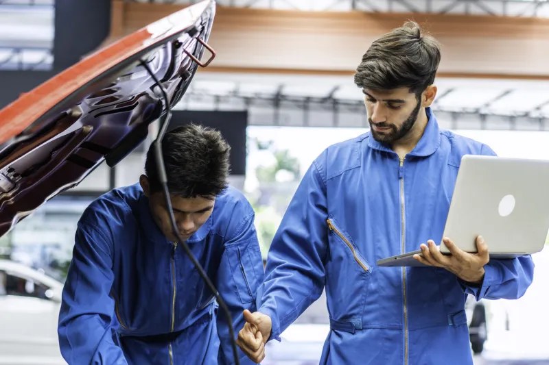 caucasian automobile mechanic man using laptop computer diagnostic and repairing car while his colleague checking in damage broken part at garage automotive, motor technician maintenance after service