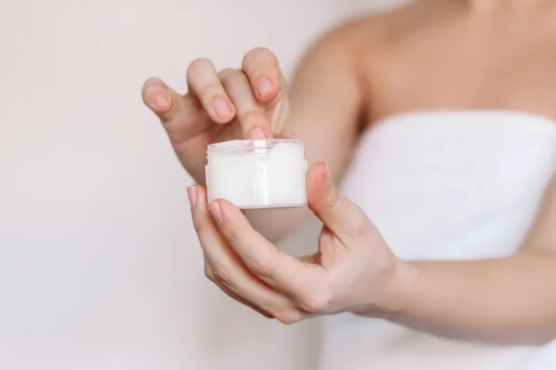 cropped shot of a young caucasian woman in a white towel after shower taking moisturizing cream with her finger holding cosmetic jar in her hand on a light background skin care