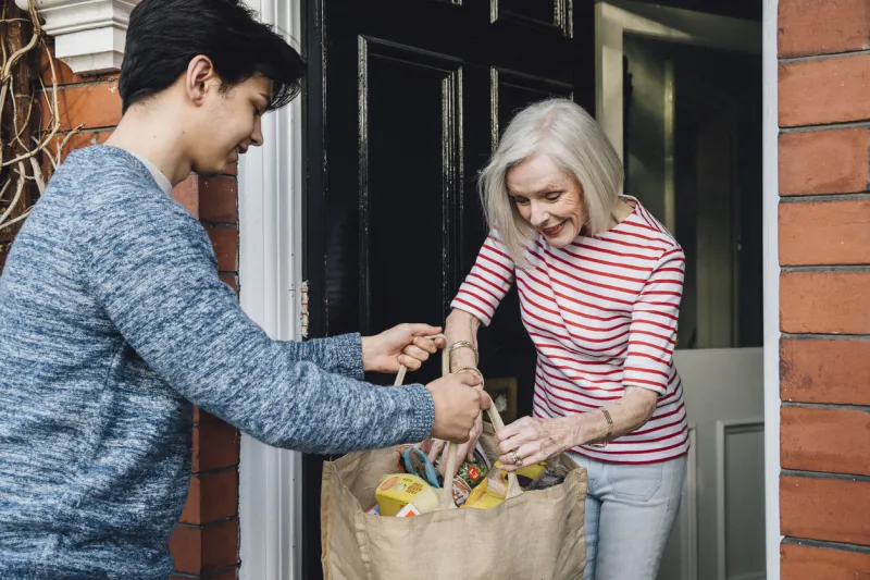 teenage boy is delivering some groceries to an elderly woman he is handing her a shopping bag at her front door