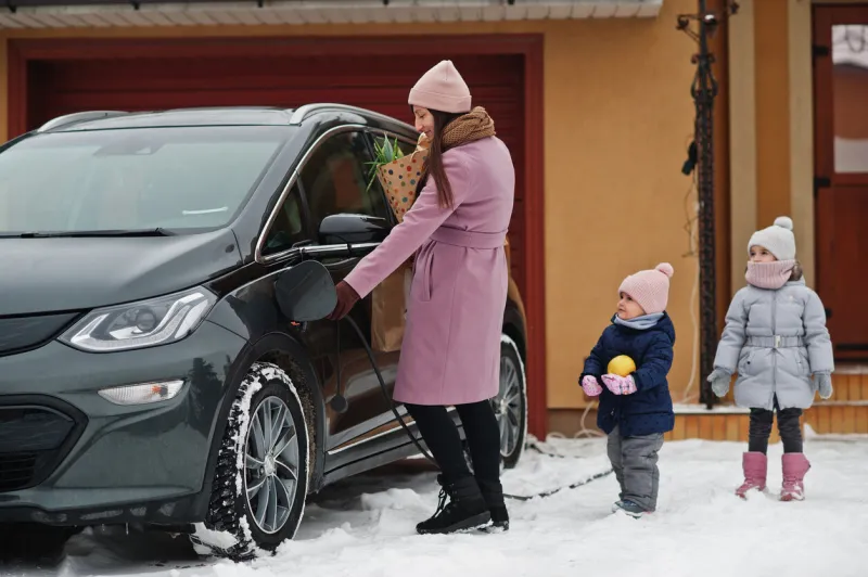 young woman with kids hold eco bags and charging electric car in the yard of her house