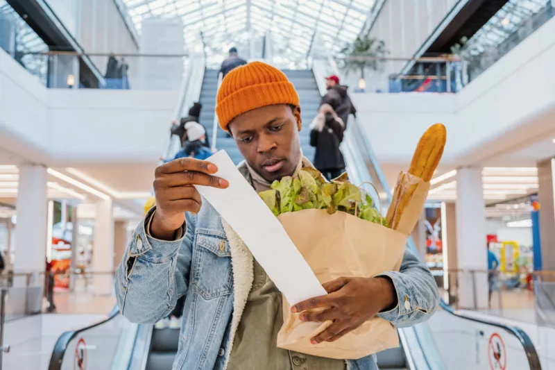 surprised african-american man in denim jacket looks at receipt total in sales check holding paper bag with products in mall