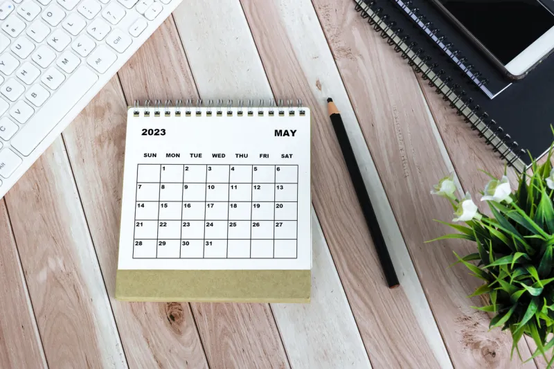 white may 2023 calendar on wooden desk with keyboard, note books, pencil, potted plant and smartphone