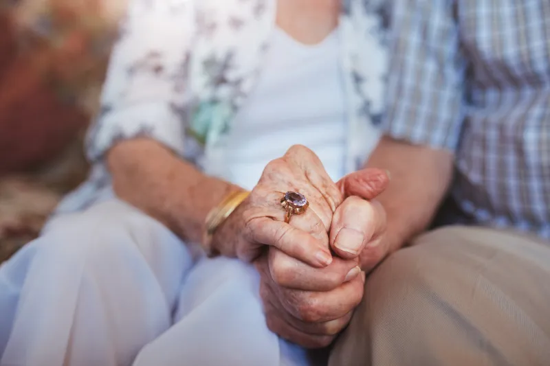 cropped shot of elderly couple holding hands while sitting together at home focus on hands