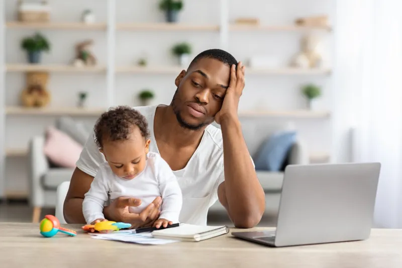 tired african american father with infant baby working on laptop at home office, exhausted black freelancer man sitting at desk with computer and holding little child, having multitasking stress