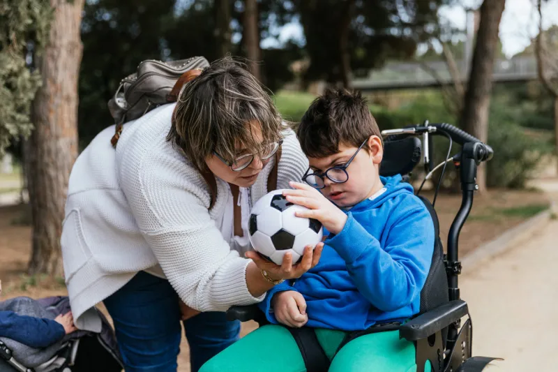 child with multiple disabilities in a wheelchair playing with a soccer ball with her mother outdoors disabled people and childhood concept