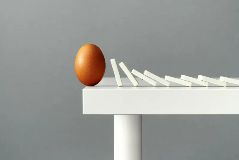 egg balancing on the edge of a table, about to fall down and break due to domino tiles falling