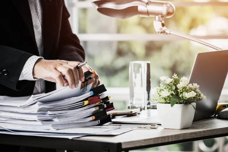 businessman hands holding pen for working in stacks of paper files searching information business report papers and piles of unfinished documents achieves on laptop computer desk in modern office