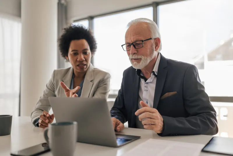 female executive discussing with senior professional while working on laptop at desk in office