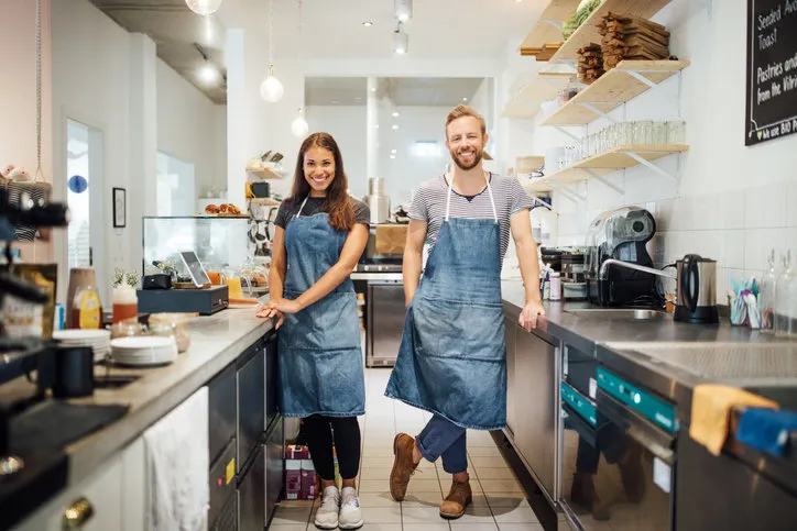 two young multiracial baristas in aprons standing in kitchen of coffee shop smiling man and woman standing by kitchen counter of cafe and looking at camera