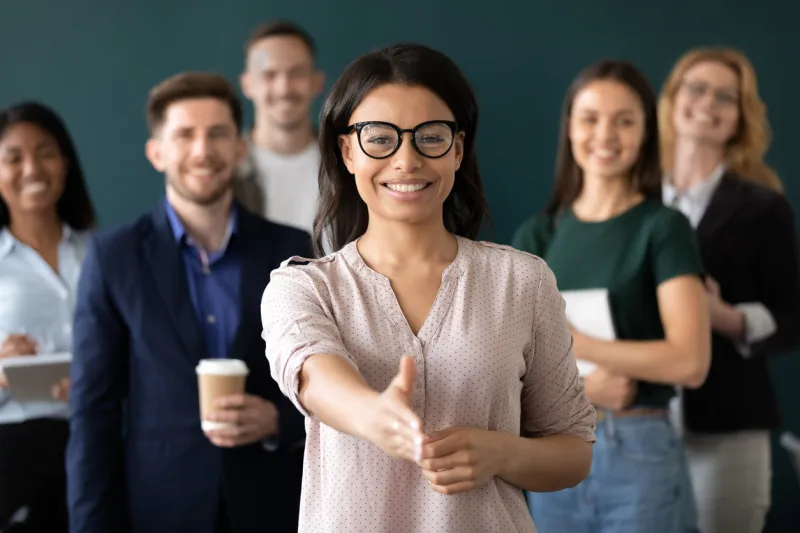 mixed race woman sales manager stretch out hand introduces herself greeting shake hands client smiling look at camera pose indoors with diverse teammates hr, job interview, business etiquette concept