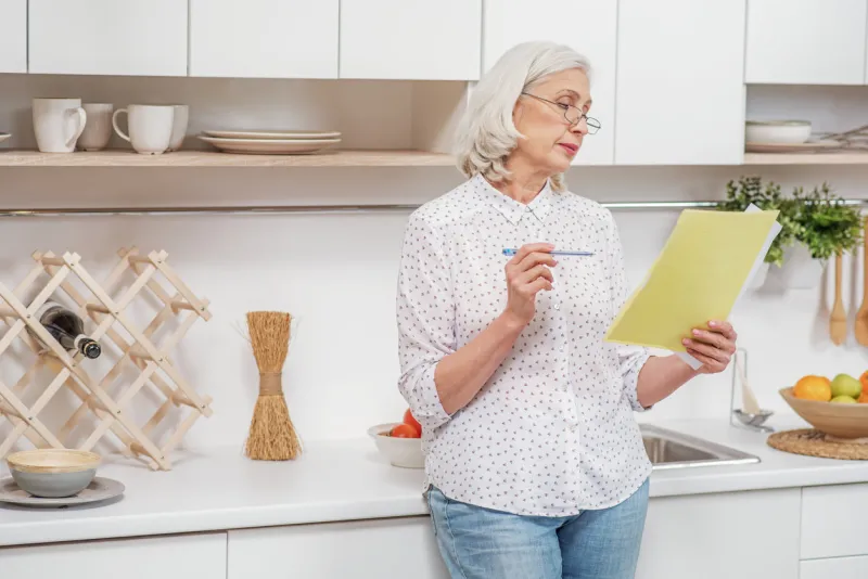 pensive senior housewife is signing insurance contract she is standing in domestic kitchen