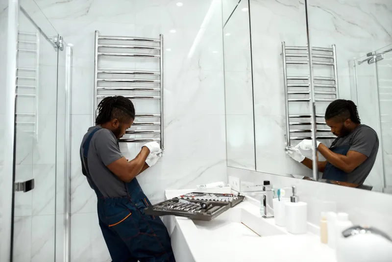 portrait of handsome mixed-race plumber in overalls and white gloves, fixing electric towel rail in modern bathroom with large mirror opened toolkit placed near on sink mirror reflection