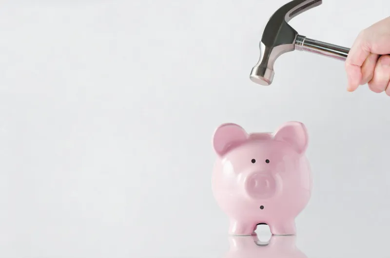 a hand holding a hammer which is raised above a pink china piggy bank, with a shocked and apprehensive facial expression reflective surface and light grey background copy space to the left