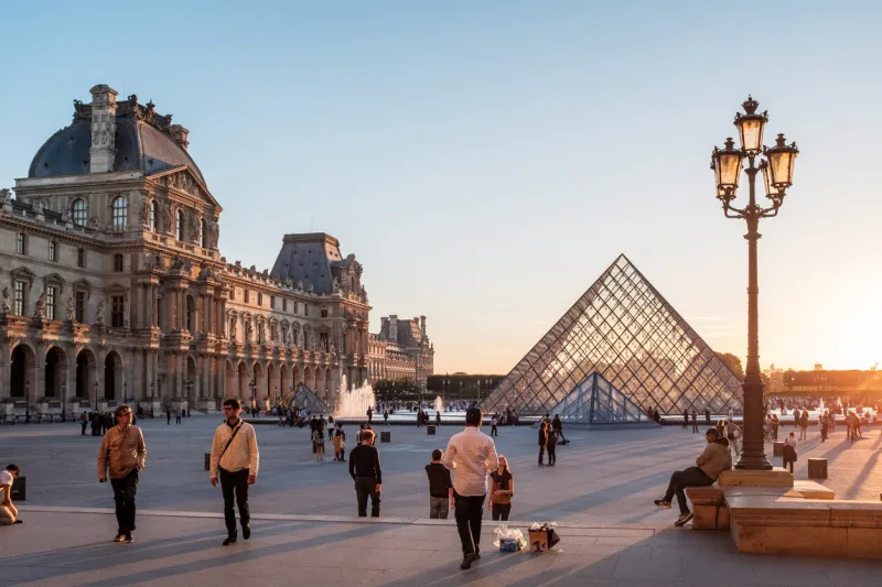 paris, france, june 22, 2018  the sun begins to rise at an already busy louvre, casting a beautiful glow against the pyramids, the lamp post, and the facade of the museum