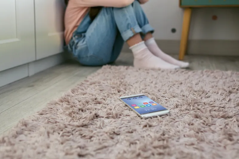 mobile phone lying on the floor with unrecognizable sad little girl sitting behind selective focus on mobile in foreground