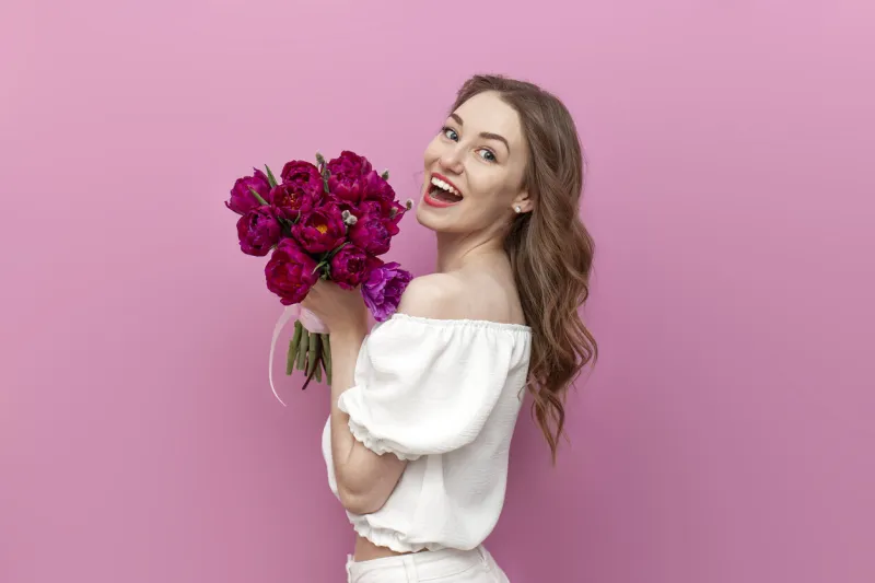 young attractive girl in white clothes holds bouquet of pink tulips and smiles, woman on holiday with flowers on pink isolated background, concept of spring and march