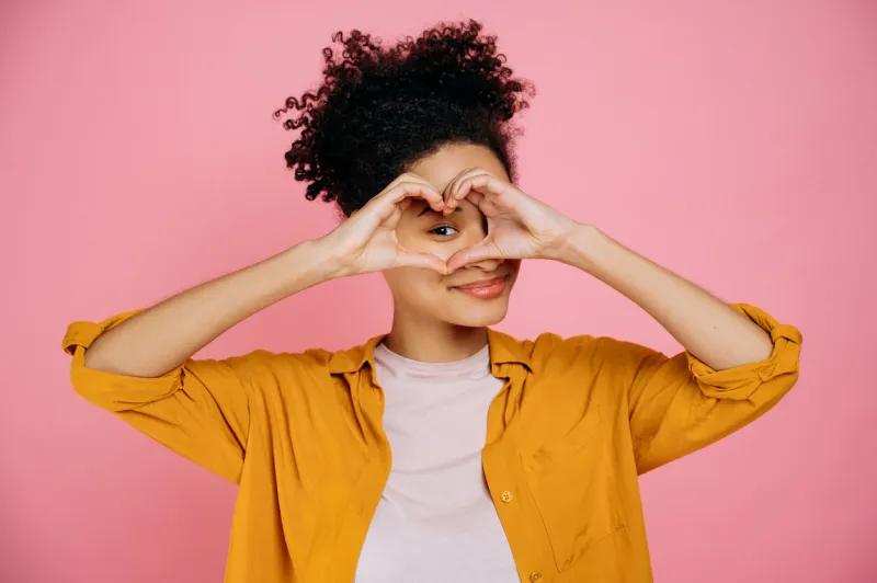 lovely candid pleasant african american girl, makes heart gesture with hands near eye, demonstrates love sign, stands on isolated pink background, looking at camera though love gesture, smile friendly