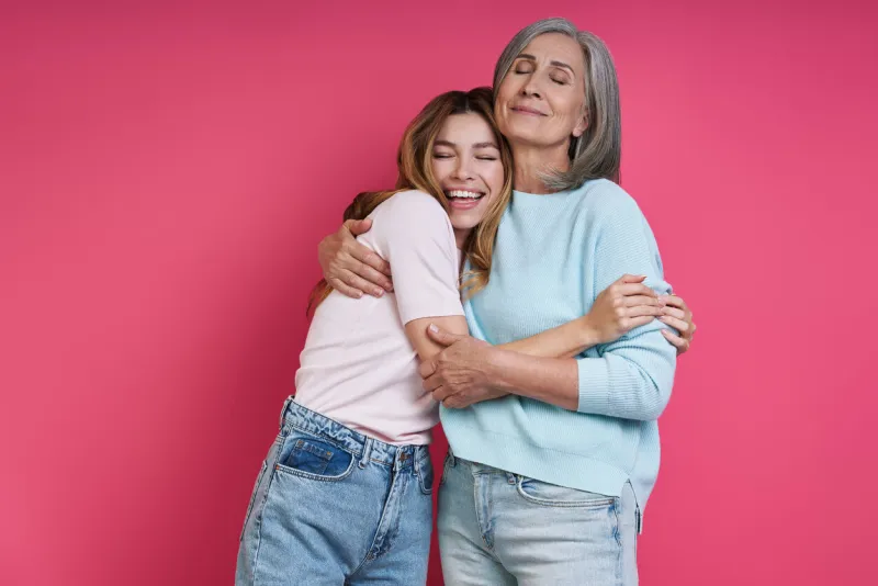 happy mother and adult daughter embracing against pink background