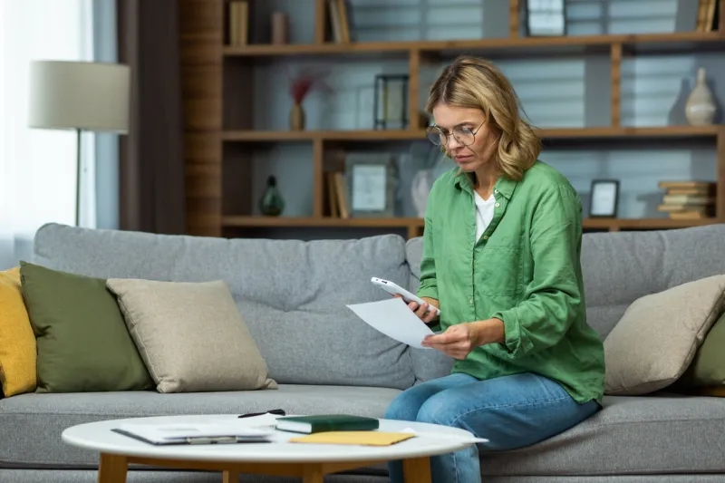 serious and confused senior woman in glasses sitting on sofa at home worriedly holding the phone and documents in his hands problem with accounts, credit, payment