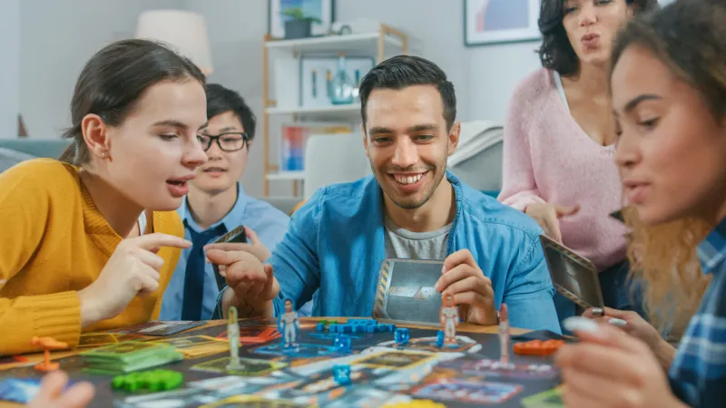 diverse group of guys and girls playing in a strategic uniquely designed board game with cards and dice friends having fun reading cards, joking, making moves and laughing in a cozy living room