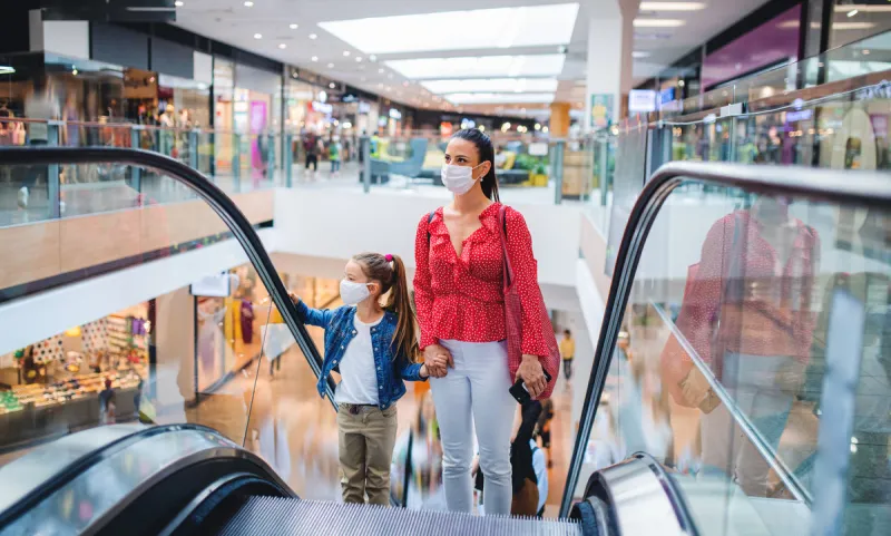 young mother and daughter with face mask on escalator indoors in shopping center, coronavirus concept