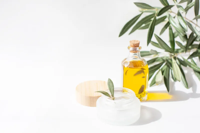 a glass jar with olive oil and olive tree twig are on white background natural cosmetics theme copy space