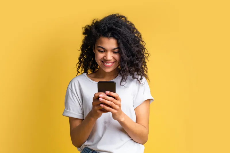 cute happy blogger girl with perfect smile in basic white t-shirt using her mobile phone answering to her followers, texting to her boyfriend, browsing various applcations device addiction concept
