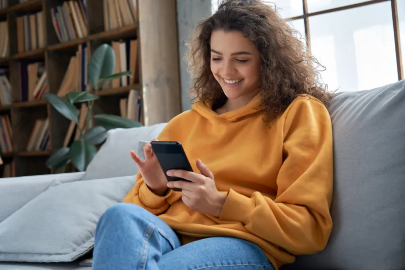 happy millennial hispanic teen girl checking social media holding smartphone at home smiling young latin woman using mobile phone app playing game, shopping online, ordering delivery relax on sofa